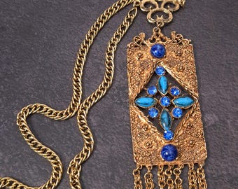 May include: A gold-tone metal pendant necklace featuring a rectangular filigree frame with a central diamond-shaped cluster of blue rhinestones and turquoise-coloured cabochons. Two dark blue round stones accent the top and bottom, with chain fringe.
