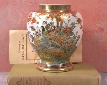 May include: A white ceramic vase with a gold rim and base, featuring a detailed hand-painted scene of a pheasant among colourful flowers and foliage. The vase is positioned in front of a paper booklet titled The story of SATSUMA WARE.