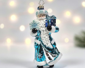May include: A glass Christmas ornament shaped like a figure with a long white beard, wearing a metallic teal coat with white snowflake patterns and white trim. The figure holds a large blue gift box and a snowflake, topped with a star hanger.