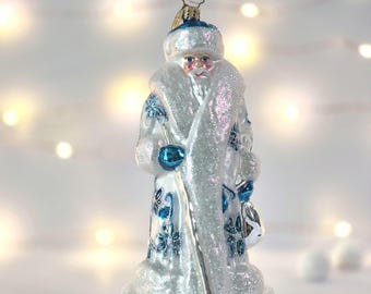 May include: A glass Christmas ornament shaped like a figure with a long white beard, wearing a long white coat covered in silver glitter and decorated with blue floral patterns. The figure wears a matching hat and holds a small blue bag.