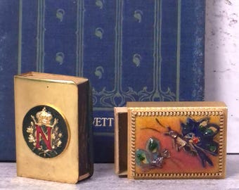 May include: Two rectangular metal matchbox covers. One is gold-toned with a circular emblem featuring a red letter N, a crown, and green enamel. The other is gold-toned with a textured border and a colourful enamel inlay depicting a butterfly and insect.