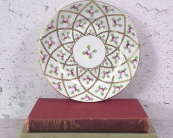 May include: A round porcelain saucer featuring a white centre with a small pink rose and green leaf motif, surrounded by a gold lattice pattern containing additional pink rose and green leaf accents throughout the entire surface area.