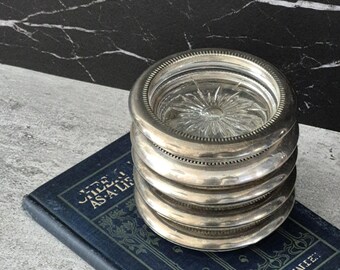May include: A stack of five circular coasters featuring silver-toned metal rims with a beaded edge and clear glass centres with a starburst pattern. The coasters are resting on a dark blue book with gold text reading "STEEL AS A LIFE".