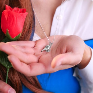 May include: A silver necklace with a small, detailed charm of a genie lamp. The lamp is blue and gold and has a swirl design. The necklace is worn by a person holding a red rose.
