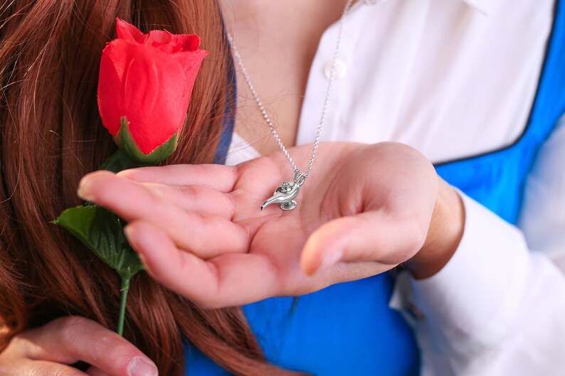 May include: A silver necklace with a small, detailed charm of a genie lamp. The lamp is blue and gold and has a swirl design. The necklace is worn by a person holding a red rose.