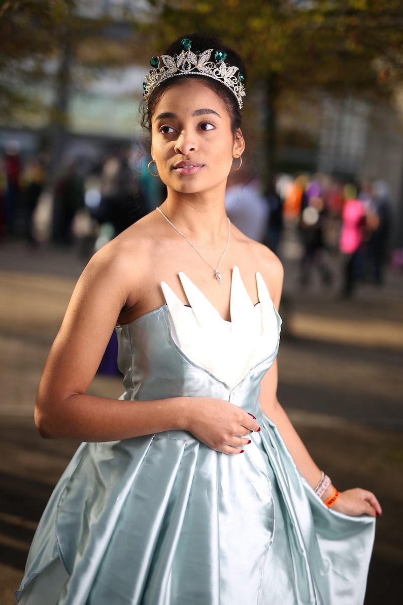 May include: A woman wearing a light blue satin dress with a white bodice and a green jeweled crown. She is standing in front of a blurred background.