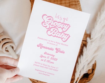 May include: A white baby shower invitation with pink text that reads "Let's get Groovy Baby." The invitation includes the name "Amanda Gale," the date, time, and RSVP information. A wooden rattle and dried plant are in the background.