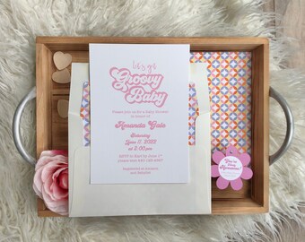 May include: A baby shower invitation with the text "Let's get Groovy Baby" in pink script. The invitation is in a cream envelope, inside a wooden tray with a floral accent and a flower-shaped tag that says "You're so awesome!"