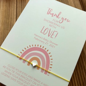May include: A baby shower invitation card with a yellow string bracelet and a small silver heart charm. The card features a pink rainbow design and the text "Thank you for showering us with love!"