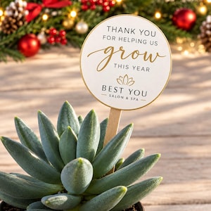 May include: A green succulent plant in a dark pot with a round wooden sign on a stick. The sign features gold and black text that reads: THANK YOU FOR HELPING US grow THIS YEAR BEST YOU SALON & SPA with a small lotus flower icon.