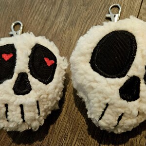May include: Two plush skull-shaped keychains. Each keychain features a white, fluffy exterior with black eyes and nose, and a silver metal clip. One skull has red heart-shaped accents in the eyes.