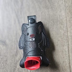 May include: Black faux leather  inhaler case with an embroidered bat design. The case has a red button and a red inhaler holder.