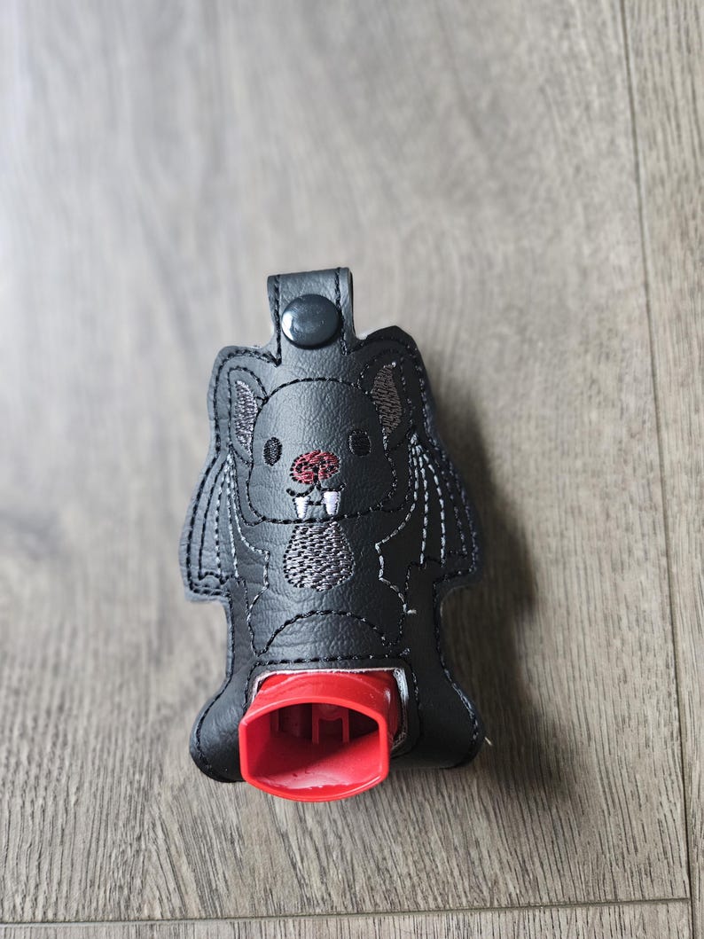 May include: Black faux leather  inhaler case with an embroidered bat design. The case has a red button and a red inhaler holder.