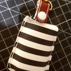 May include: A black and white striped fabric pouch with a brown leather strap and a gold metal clip. The pouch has a white button on the strap.