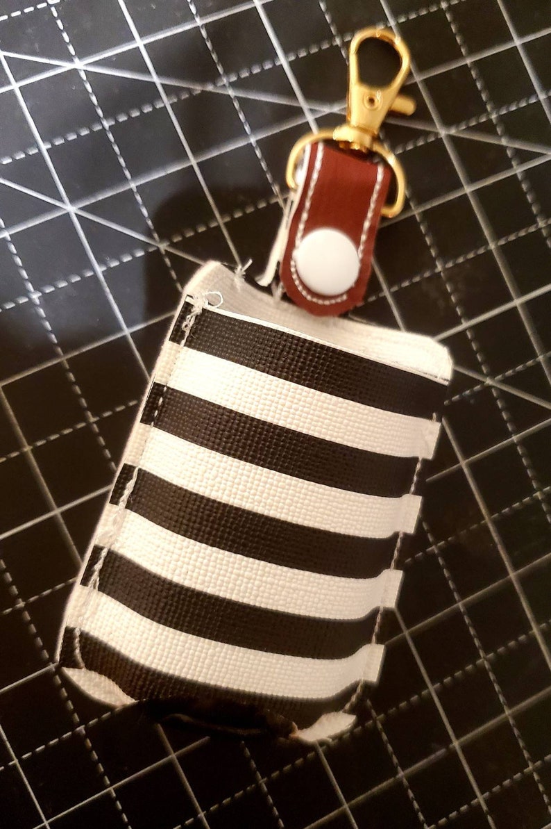May include: A black and white striped fabric pouch with a brown leather strap and a gold metal clip. The pouch has a white button on the strap.