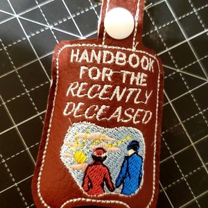 May include: A brown leather hand sanitizer holder with white stitching. The holder has a white button and a loop for attaching to a keychain. The front of the holder has white embroidery that reads "Handbook for the Recently Deceased" and features a cartoon image of two people walking away from a yellow sun.