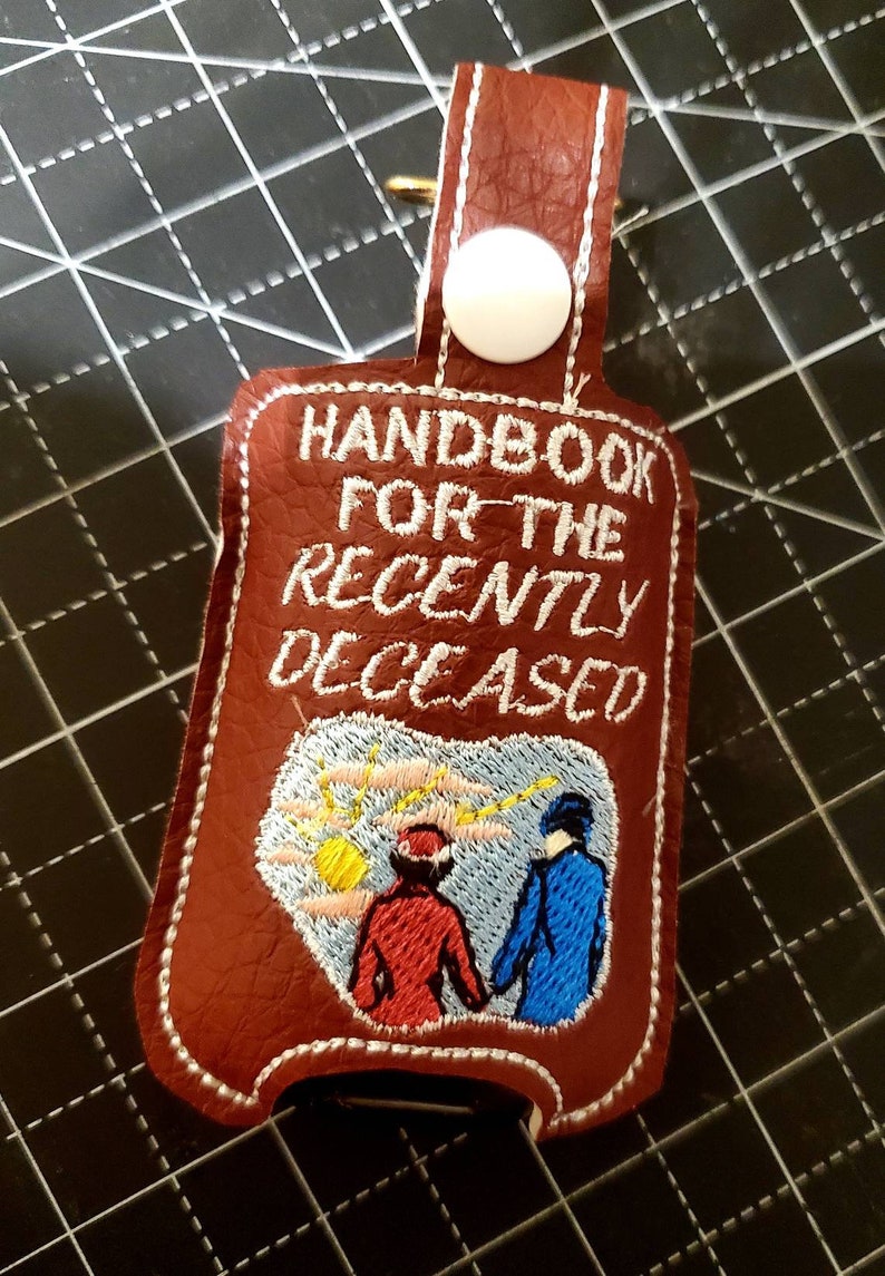 May include: A brown leather hand sanitizer holder with white stitching. The holder has a white button and a loop for attaching to a keychain. The front of the holder has white embroidery that reads "Handbook for the Recently Deceased" and features a cartoon image of two people walking away from a yellow sun.