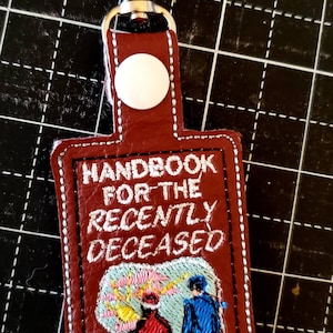 Może przedstawiać: Brązowy skórzany brelok z białym haftem. Haft przedstawia tekst "Handbook for the Recently Deceased" i kreskówkowy obrazek dwóch osób odchodzących od jasnego światła.