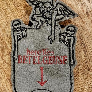 May include: A gray felt keychain with a black embroidered image of a skeleton with wings and the text "here lies BETELGEUSE" in red. The keychain has a silver metal clasp.