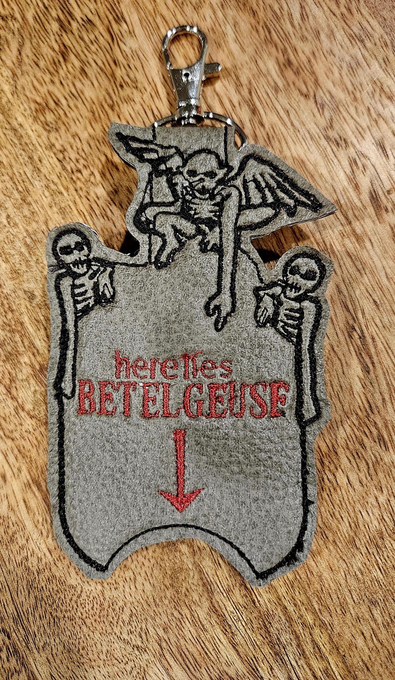 May include: A gray felt keychain with a black embroidered image of a skeleton with wings and the text "here lies BETELGEUSE" in red. The keychain has a silver metal clasp.