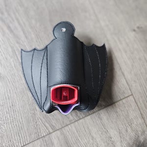 May include: Black bat-shaped fabric holder for a small spray bottle. The holder has a purple lining and the bottle has a red top.
