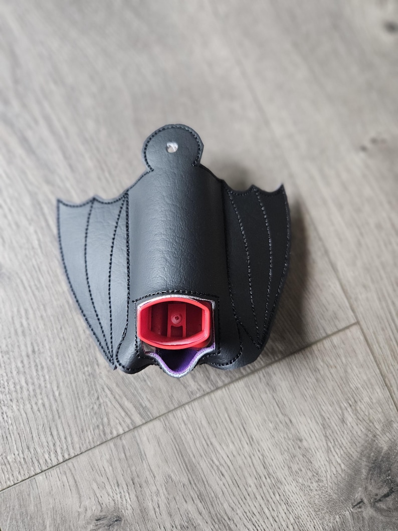 May include: Black bat-shaped fabric holder for a small spray bottle. The holder has a purple lining and the bottle has a red top.