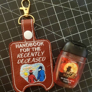 May include: A brown leather hand sanitizer holder with the text "HANDBOOK FOR THE RECENTLY DECEASED" and an embroidered image of two people walking. A small red bottle of hand sanitizer with the text "VAMPIRE BLOOD" is next to the holder.