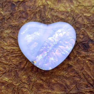 May include: A heart-shaped gemstone with a pale blue hue and iridescent internal patterns. The stone has a polished surface and is set against a textured brown background. The gemstone is likely an opal.