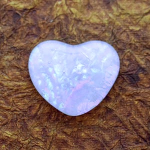May include: A heart-shaped, iridescent gemstone with a pale lavender hue. The stone has a polished surface and displays flashes of pink and blue. The background is a textured, brown surface.