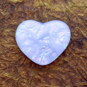 May include: A heart-shaped, iridescent, light blue stone with a shimmering, textured surface. The stone has a pearlescent quality and is set against a textured, brown background. The heart shape is clearly defined.