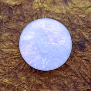 May include: A round, opalescent gemstone with a swirling, textured pattern. The gem has a soft, pearly white color with hints of pink and blue. The stone is set against a textured, brown background.