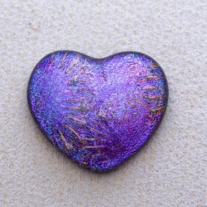 May include: A dichroic glass heart in shades of purple, blue, and gold. The heart-shaped pendant has a textured surface that reflects light, creating a shimmering effect. The heart is a decorative item.