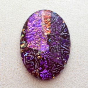 May include: Oval-shaped dichroic glass cabochon with a vibrant purple and iridescent color scheme. The glass features a textured surface with intricate patterns and a mix of purple, gold, and orange hues. This cabochon is suitable for jewelry making.