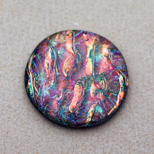 May include: A circular glass cabochon featuring a vibrant, iridescent finish with swirling patterns of deep purple, magenta, teal, and gold. The surface displays a reflective, metallic sheen that shifts in color across the rounded, smooth texture.
