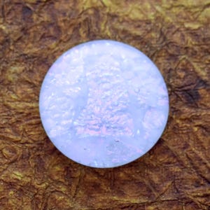 May include: A round, iridescent glass cabochon with a light blue and pink color scheme. The cabochon has a textured surface and is suitable for jewelry making or other craft projects.