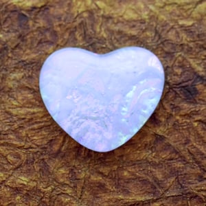 May include: A heart-shaped, iridescent glass object with a pale blue and white color scheme. The glass has a textured appearance, and the heart is centered on a brown, textured surface. The object is likely a decorative item.