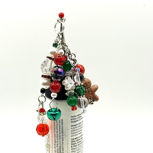 May include: A Christmas-themed charm with a variety of beads in red, green, white, and iridescent colors. The charm includes a small gingerbread man, a green bell, and a red bead at the top. The charm is attached to a silver chain.