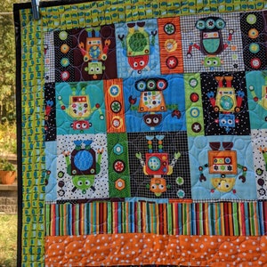 Könnte beinhalten: Eine farbenfrohe Quiltdecke mit Roboter-Motiv. Die Decke besteht aus verschiedenen Stoffen mit unterschiedlichen Mustern, darunter Streifen, Punkte und geometrische Formen. Die Roboter sind in verschiedenen Farben und Designs dargestellt.