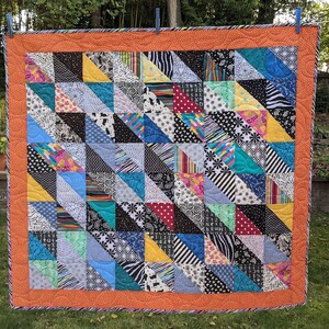 Może przedstawiać: Kołdra w patchworku z różnorodnych kolorowych tkanin. Kołdra składa się z trójkątów w różnych kolorach i wzorach, w tym czarnym, białym, niebieskim, czerwonym, zielonym i żółtym. Kołdra jest obszyta jasno pomarańczową lamówką.