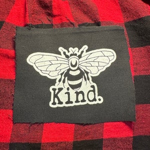 May include: A black fabric patch with a white bee design and the word "Kind." sewn onto a red and black plaid fabric.