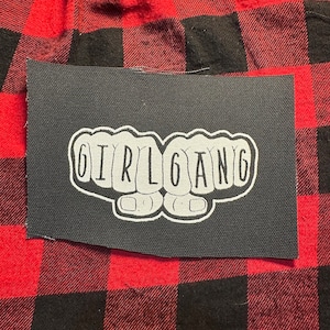 Puede incluir: Parche de plancha negro con texto blanco que dice "GIRL GANG" en una fuente estilizada. El texto está formado por dos puños con los dedos deletreando las palabras.