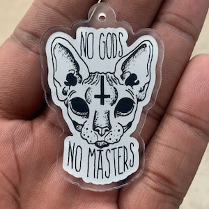 Pode incluir: Um chaveiro de acrílico transparente com uma ilustração em preto e branco de um gato Sphynx com uma cruz na testa. O texto "NO GODS NO MASTERS" está impresso abaixo do gato.