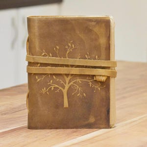 Puede incluir: Un diario de cuero marrón con un diseño de árbol en relieve en la portada. El diario tiene dos correas de cuero que se envuelven alrededor de la portada y se atan en la parte delantera.