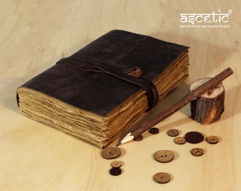 May include: A brown leather-bound journal with a leather strap closure, a pencil, and wooden buttons on a wooden surface. The journal has a rustic, handmade look. The text "Ascetic World of Artistic Paper & Leather Products" is visible in the top right corner of the image.