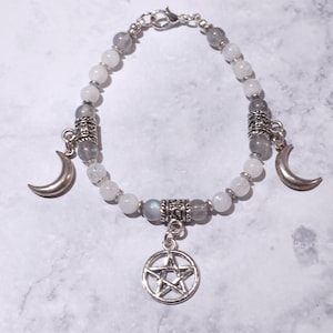 May include: A beaded bracelet featuring alternating white and translucent gray round beads, silver-toned metal spacers, two silver crescent moon charms, and a central silver pentacle charm. The bracelet is secured with a silver-toned lobster claw clasp.