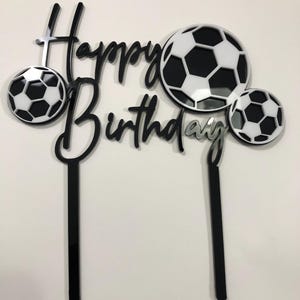 May include: A black and silver cake topper with the words "Happy Birthday" and three soccer balls. The soccer balls are white with black pentagon shapes. The topper has two black stakes for insertion into a cake.
