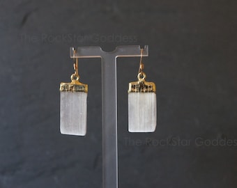 Pode incluir: Um par de brincos de selenita com detalhes em metal dourado. Cada brinco apresenta uma pedra retangular de selenita branca com uma tampa dourada e um gancho dourado. Os brincos são exibidos em um suporte transparente sobre um fundo escuro.