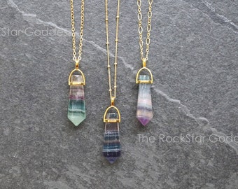 May include: Three gold necklaces with rainbow fluorite crystal pendants. The necklaces are layered and the crystals are different sizes. The necklaces are on a gray background.