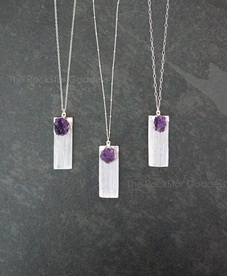 May include: Three silver necklaces with selenite crystal pendants and amethyst crystal accents. The necklaces are on a dark gray background.