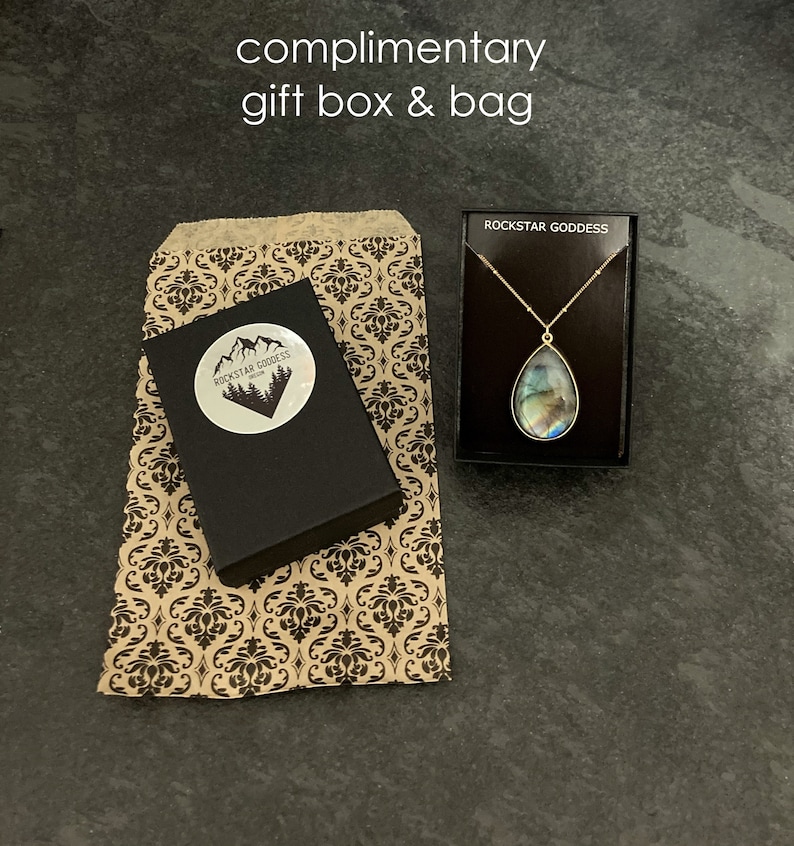 Puede incluir: Una bolsa de regalo de papel marr&oacute;n con un patr&oacute;n damasco en blanco y negro. La bolsa est&aacute; al lado de una caja de regalo negra con una pegatina blanca que dice "Rockstar Goddess Jewelry".  Tambi&eacute;n se muestra una peque&ntilde;a caja negra con un collar de cadena dorada y un colgante azul iridiscente. La caja tiene el texto "Rockstar Goddess" impreso en la parte superior.